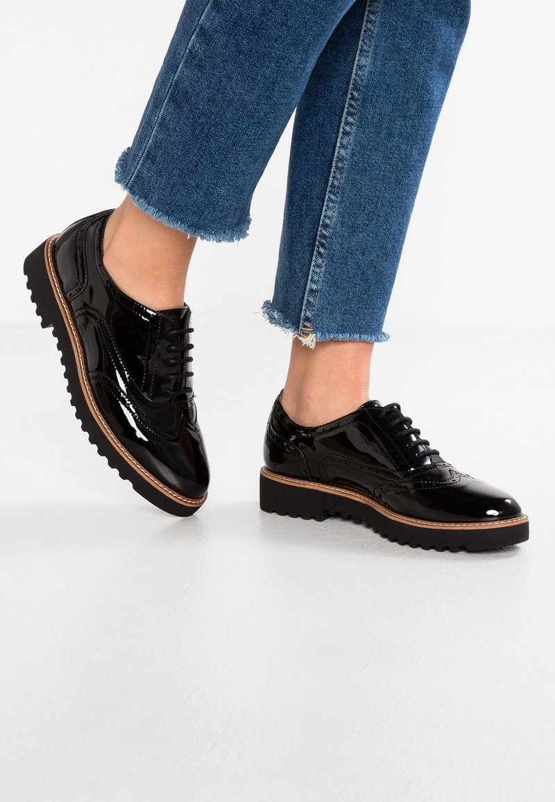 Des chaussures en cuir verni noir présentent un design brogue classique avec fermeture à lacets et une semelle épaisse noire, accentuée d'un bord marron contrastant.