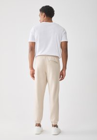 Mann sett fra bak som har på seg en hvit t-skjorte, beige joggebukser med baklomme, og hvite sneakers mot en ensfarget hvit bakgrunn.