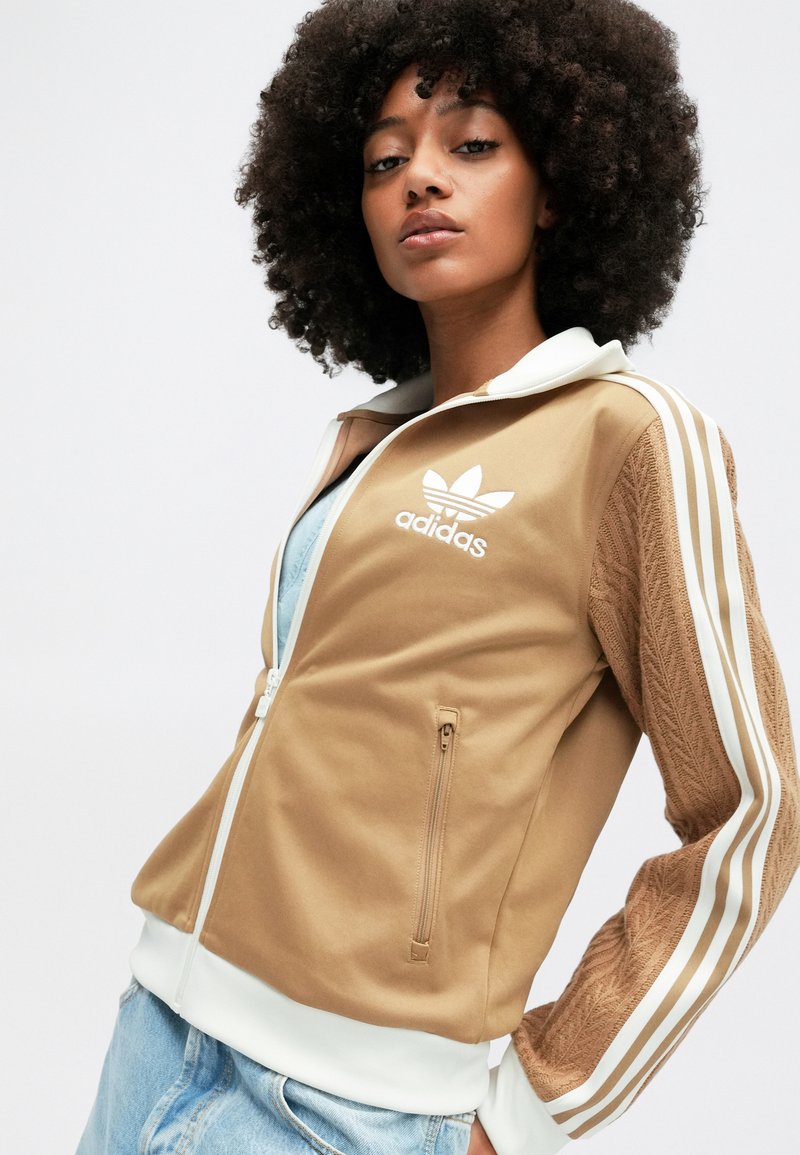 Braune Sportjacke mit weißen Akzenten, Reißverschluss, strukturierten Ärmeln und ikonischem Logo auf der Brust. Kombiniert mit einem hellblauen Oberteil und Jeans.