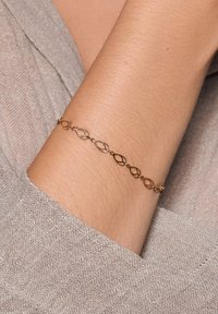 Złocista bransoletka z połączonymi owalnymi kształtami oraz małymi okrągłymi koralikami pomiędzy. Noszona na nagim nadgarstku na tle fakturowanej tkaniny.