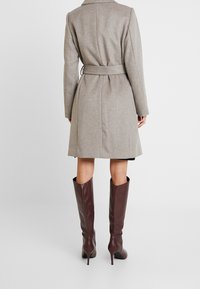 Abrigo de mezcla de lana beige con cintura ceñida y mangas largas, combinado con botas de cuero burdeos de tacón hasta la rodilla. Textura suave y ajuste entallado.