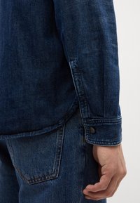 Tumši zila džinsu krekls ar pogām pie aproces, ar smalku tekstūru un izliekta apakša. Saskaņota ar atbilstošām zilām džinsa biksēm.