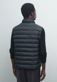 Homme portant un gilet matelassé noir et une chemise à manches longues, se tenant tourné vers le dos sur un fond gris clair uni.