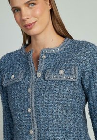 Giacca in tweed blu con texture, caratterizzata da un collo rotondo, dettagli in bottoni argentati e due tasche frontali con decorazioni in paillettes.