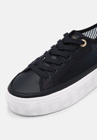Černá textilní teniska s texturovaným designem, hladkou koženou špičkou, černými tkaničkami a bílou vzorovanou gumovou podrážkou.