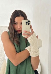 Roomkleurige, gebreide wanten zonder vingers met geribbelde manchetten, gedragen door een persoon, gecombineerd met een groene mouwloze top. Zachte textuur en gezellig ontwerp.