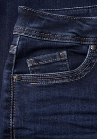 Jean en denim bleu foncé avec surpiqûres et une petite poche avant. Le matériau semble doux avec une légère texture. Détails de bouton et de rivets visibles.