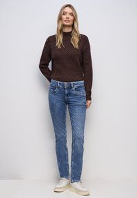 Žena stojící u bílé zdi, oblečená v tmavě hnědém svetr s copánkovým vzorem, modrých džínách a bílých teniskách s béžovými podrážkami.