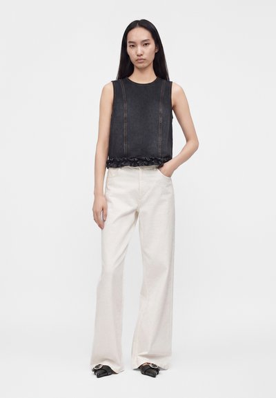 Jeune femme aux longs cheveux noirs portant un top noir sans manches avec un ourlet à volants et un pantalon blanc large, debout avec une main dans la poche.