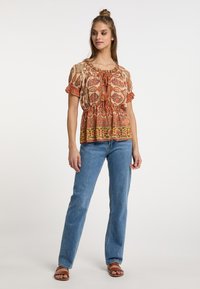 Blusa con motivo e scollo arricciato, maniche corte a palloncino e un accento in vita, in calde tonalità di arancione e crema, abbinata a jeans azzurri chiari.