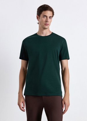 Mann steht vor weißem Hintergrund, trägt ein dunkelgrünes kurzärmeliges T-Shirt und braune Hose, schaut leicht zur Seite.