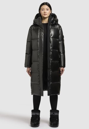 Woman wearing a long, shiny black puffer coat with hood, black leggings, and black winter boots standing against a plain light background.