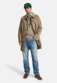 Mann in grüner Mütze, beigem Trenchcoat, grauem Pullover, hellblauen Jeans und braunen Lederschuhen, der mit einer Hand in der Tasche steht.