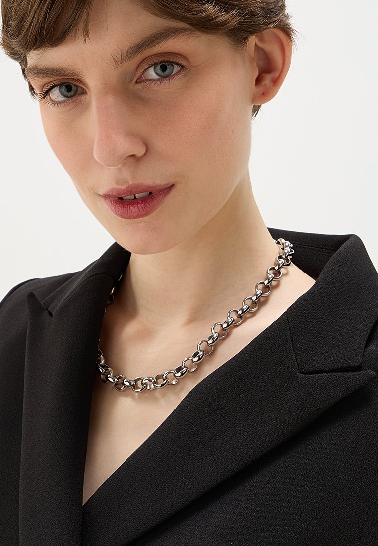 Collana in argento con grandi anelli intrecciati. Il modello indossa un blazer nero, mettendo in risalto la collana attorno al collo.