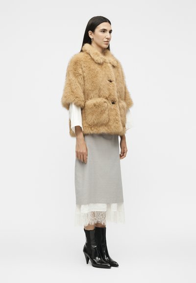 Femme portant une cape en fourrure beige, un haut blanc à manches longues, une jupe midi grise avec un ourlet en dentelle et des bottines noires à talons, debout devant un fond uni.