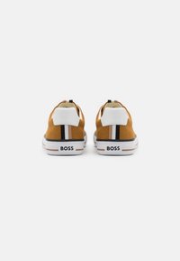 Par de zapatillas de lona color mostaza con parches blancos en el talón, rayas negras y beiges, y la marca "BOSS" en las suelas blancas, vistas desde atrás.
