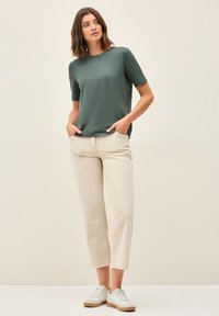 Mørkegrøn kortærmet t-shirt med afslappet pasform, parret med lysebeige bukser. Modellen har hvide sneakers på med en lys tan sål.