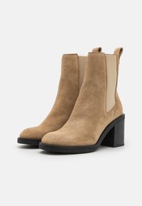 Botas de tobillo de ante en color marrón claro con paneles laterales elásticos, punta redondeada y un tacón grueso negro. Superficie texturizada con un acabado suave.