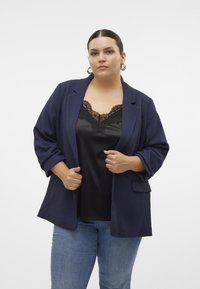 Navy blazer with a textured finish, notched lapels, side pockets, and relaxed fit, layered over a black satin tank top with lace trim.