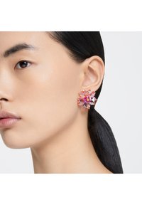 Brinco em forma de flor de cristal rosa cintilante com múltiplas facetas, detalhes em tom dourado e uma gema vermelha central, usado na orelha de um modelo.