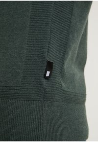 Dunkelgrüner Strickpullover mit gerippter Textur und kontrastierendem glattem Einsatz. Verfügt über ein kleines schwarzes Etikett an der Seitennaht.