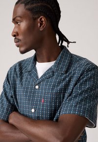 Korte mouwen shirt in donker teal stof, met een geometrisch patroon in crème en roest, voorzien van een notch-kraag en een merklabel nabij de zak.