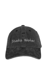 Crna traper kapa s zakrivljenim vizirjem, s bijelim izvezenim tekstom "Studio Worker" na prednjoj strani. Šestopanelna konstrukcija s ventilacijskim rupama.