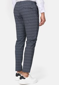 Schmal geschnittene, marineblaue karierte Hose mit einem strukturierten Muster und schmalem Bein. Getragen mit weißen Sneakers. Mit einer einzelnen Rückentasche.