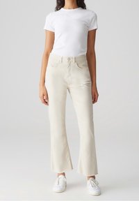 Femme portant un t-shirt blanc ajusté, un jean droit écru avec des ourlets bruts, et des baskets blanches, debout devant un fond uni.