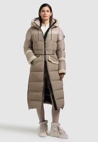 Cappotto lungo imbottito di un beige chiaro con cappuccio foderato in pelliccia, chiusura a zip, due tasche laterali e una cintura. Indossato con leggings chiari e stivali.
