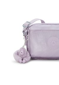 Lilafarbene Handtasche mit glatter Oberfläche, ausgestattet mit einem runden Logo, verstellbarem Riemen und einem plüschigen Affen-Schlüsselanhänger, der mit einem Metallclip befestigt ist.