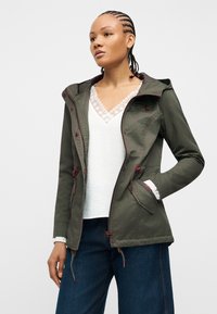 Femme aux cheveux tressés portant une veste à capuche vert olive sur un chemisier blanc orné de dentelle et un jean large bleu foncé, les mains dans les poches.