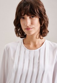 Blusa bianca con pieghe verticali, scollatura rotonda e tessuto liscio. Il tessuto sembra leggero e fluido.