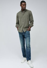Camisa de botones en verde oliva con mangas enrolladas, combinada con jeans de mezclilla azul claros y zapatillas blancas. Diseño sencillo, textura suave.