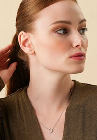 Silberne Halskette mit einem runden Anhänger, der kleine klare Steine zeigt, getragen über einem strukturierten olivgrünen Oberteil. Die Haare des Models sind nach hinten gesteckt.