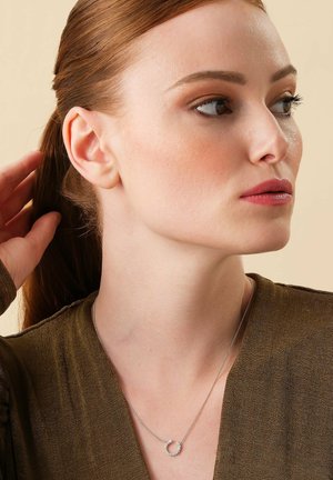 Silver necklace with a circular pendant featuring small clear stones, worn over a textured olive green top. Model's hair is pulled back.