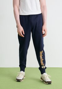 Námornícko modré teplákové nohavice s elastickým pásom a šnúrkou. Obsahujú zlaté logo "Timberland" na ľavej nohe, skombinované so svetlými teniskami.