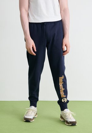 Navyblauwe joggerbroeken met een elastische tailleband en trekkoord. Voorzien van een gouden "Timberland" logo op het linkerpand, gecombineerd met lichte sneakers.