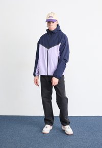 Veste coupe-vent navy et lavande avec fermeture éclair et poignets élastiques ; pantalon noir ; baskets blanches ; casquette claire avec texte.