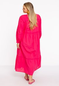 Rosa Baumwoll-Maxikleid mit langen Ärmeln, gestuftem Design und lockerer Passform. Kombiniert mit passenden rosa Rutschsandalen.