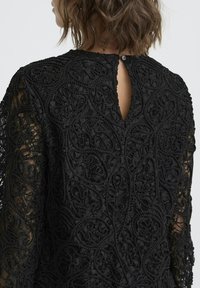 Crna čipkana bluza s složenim teksturalnim cvjetnim uzorcima, dugim rukavima i kopčanjem na dugme na leđima. Prozirni rukavi dodaju dubinu.