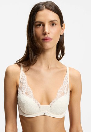Femme aux longs cheveux bruns portant un soutien-gorge en dentelle blanche avec des motifs floraux, face à l'avant sur un fond uni.