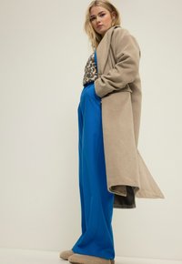 Abrigo largo beige sobre una blusa de estampado de leopardo, combinado con pantalones anchos de color azul y zapatillas beige. Tejido suave y ajuste relajado en toda la prenda.