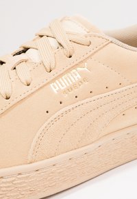 Primo piano di una sneaker Puma Suede beige in suede con suola testurizzata, lacci beige e logo Puma in rilievo sul pannello laterale.