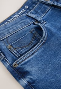 Nahaufnahme der vorderen Tasche einer blauen Denim-Jeans mit Gürtelschlaufe, Metallnieten und dem Label "MOTION FLEX" am Bund.