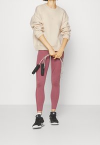 Personne portant un sweat beige, un legging mauve, des baskets noires Adidas, tenant une corde à sauter à poignée noire devant un fond uni.