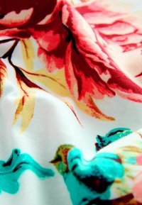 Stof met bloemmotief, gekenmerkt door grote rode en roze bloemen met gele bladeren op een witte achtergrond, aangevuld met turquoise vogels.