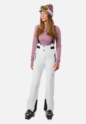 Weiße Schneehose mit schwarzen Akzenten, verstellbaren Schulterträgern und Reißverschlusstaschen. Kombiniert mit einem hellvioletten Langarmoberteil und Skibrille.