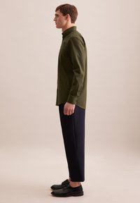 Camicia a maniche lunghe verde oliva con apertura a bottoni, tasca laterale, abbinata a pantaloni scuri e scarpe nere, mostrata di lato.