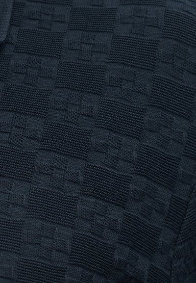 Σκοτεινό ύφασμα με υφή σε σχέδιο σκακιέρας, αποτελούμενο από εναλλασσόμενα πλεκτά τετράγωνα με ανάγλυφα γεωμετρικά σχέδια.